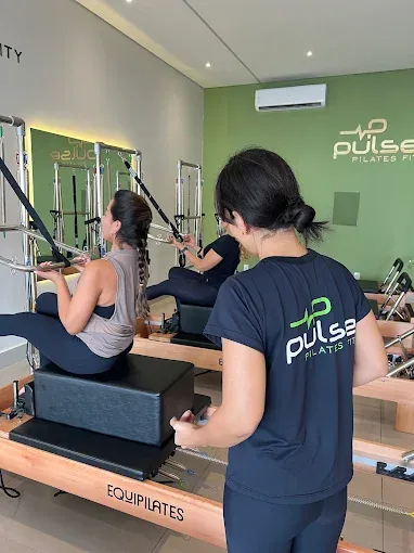 Professora orienta aluna durante aula no Pulse Pilates Fit, com aparelhos Reformer Equipilates ao fundo
